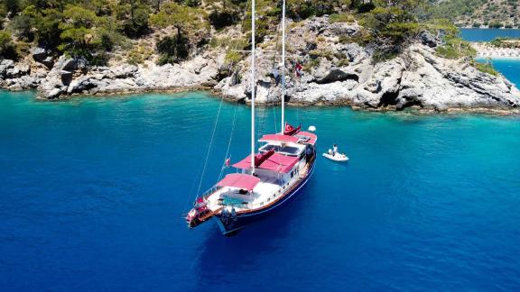 Pronto Blue - Blue gulet with red sunshades in crystal-clear waters near rocky shoreline.