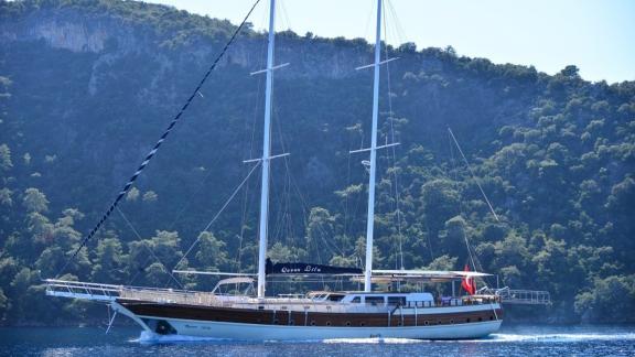 The Queen Lila sails against a backdrop of wooded hills and deep blue water.