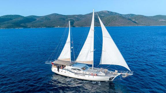 A Bodrum Gulet with 5 cabins sails on the sea in front of a hilly landscape.