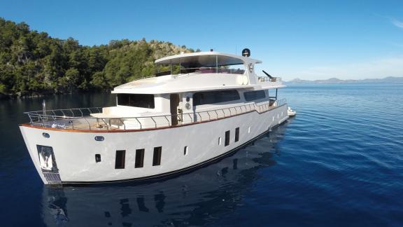 The Simay F yacht is anchored in a serene bay surrounded by lush green trees.