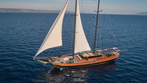 An impressive sailing yacht with white sails glides across the blue sea, with a coastline in the background.