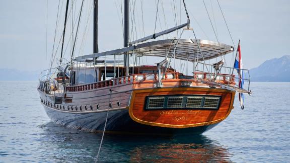 The back of an elegant sailing yacht with polished wood finish and sun deck, on a calm sea with mountains in the backgro