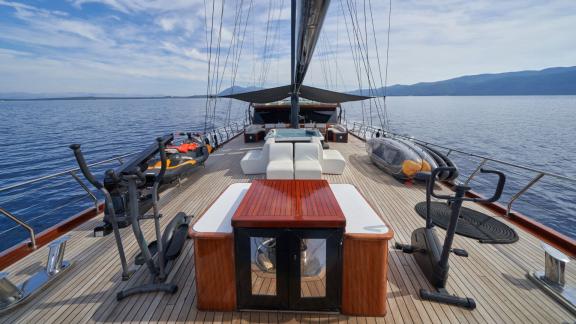 Deck of a yacht with whirlpool, fitness equipment, cosy seating and jet ski, in front of a picturesque seascape.