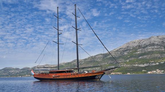 A large, elegant sailing yacht lies at anchor on calm waters, with a picturesque coastal landscape of mountains and hous