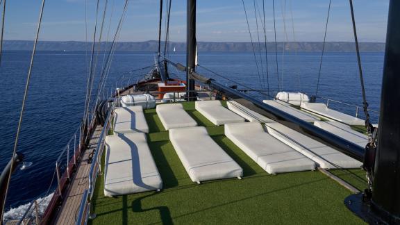 Large sun deck of a sailing yacht with white loungers on green artificial turf, surrounded by calm sea and distant mount