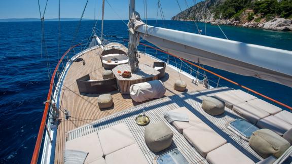 The spacious sun deck of a gulet with comfortable loungers, surrounded by azure blue water and rocky coastline.