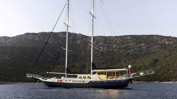 Zweimastige Segelyacht vor bewaldeten Hügeln im Wasser.