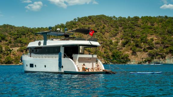 A woman relaxes at the back of the Oggusto trawler while paddleboard floats nearby in calm Fethiye bay.