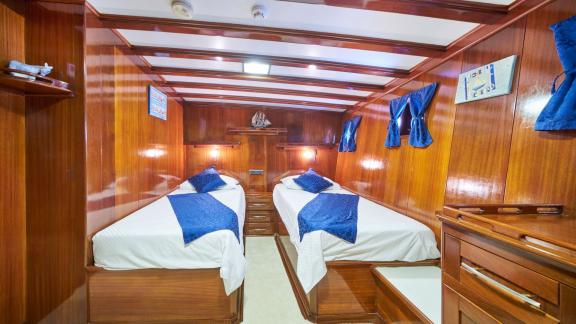 Wood-panelled cabin on a sailing yacht with two single beds, blue bed linen and small windows with curtains.