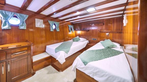 Cabin on a sailing yacht with wooden panelling, two single beds with green bed linen and small windows with curtains.