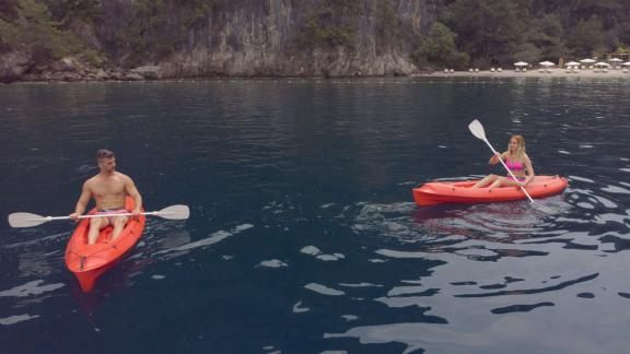 Two people enjoy kayaking on calm waters off a rocky coastline and a secluded beach.