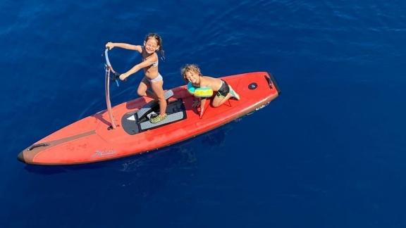 Двое детей наслаждаются прозрачными голубыми водами Гёчека (Турция), катаясь на красной паддлборде.