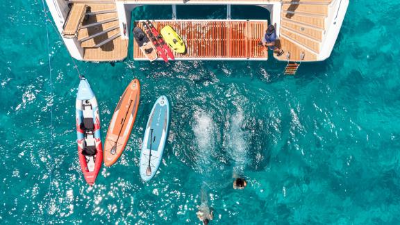 Gäste genießen Wassersportarten wie Kajakfahren und Paddleboarding neben dem Katamaran Aphaea, der in türkisfarbenem Was