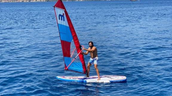 Ein Mann genießt das Windsurfen auf dem klaren blauen Wasser von Göcek, Türkei, auf einem farbenfrohen Windsurfboard.