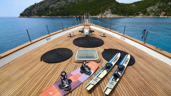 Deck eines Segelboots mit Wasserski und Wakeboard, Blick auf türkisfarbenes Wasser und bewaldete Küste.