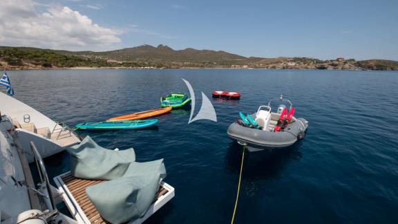 Kayaks, tender, and inflatable tubes floating behind a catamaran anchored in clear water.