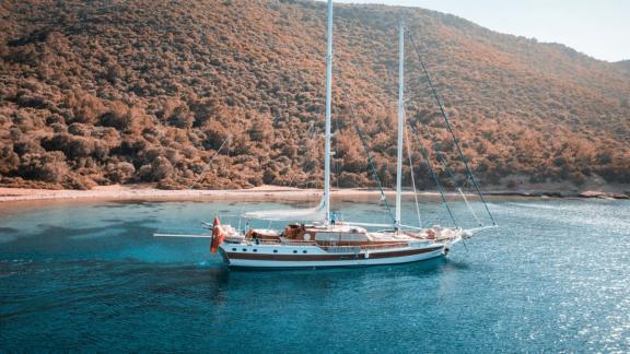 An elegant sailing ship lies quietly in a clear, turquoise-blue bay, surrounded by green hills.