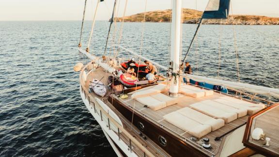 Menschen entspannen auf dem Deck eines luxuriösen Segelschiffs, umgeben von Wassersportgeräten, vor einer Küste.