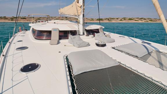 Sonnenliegen und Netze auf dem Deck eines Katamarans, vor Anker im türkisfarbenen Wasser, Küste im Hintergrund.