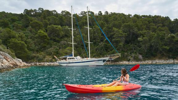 Frau im Kajak auf türkisfarbenem Wasser, im Hintergrund das Segelboot Fortuna vor bewaldeter Küste.