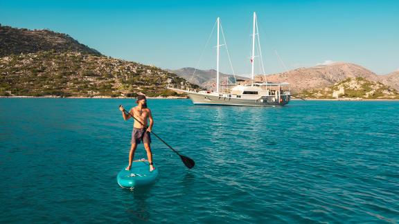 A man paddleboards on a SUP in front of the Gulet Esperanza in crystal-clear waters, surrounded by hilly landscape.