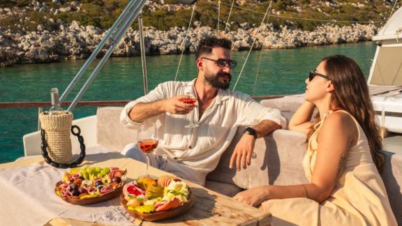 A couple relaxes with a glass of wine and a selection of snacks aboard the Gulet Esperanza, set against a rocky coastal 