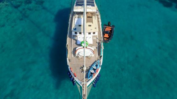 The luxury gulet Gazella is anchored in turquoise waters. The upper deck features a sunbathing area, water toys are at t