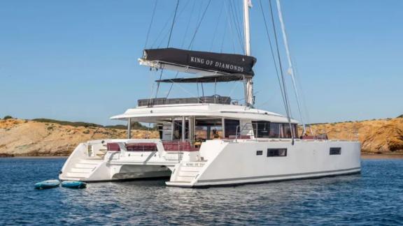 The King of Diamonds catamaran, viewed from the stern, calmly anchored near a rocky shore in Athens.