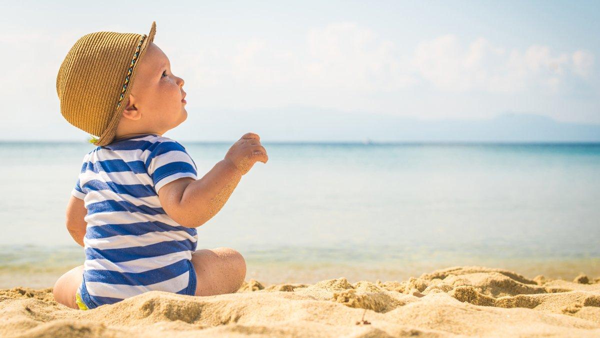 A child on the sand, in swimwear and a sun hat, happily awaits the upcoming yacht charter.