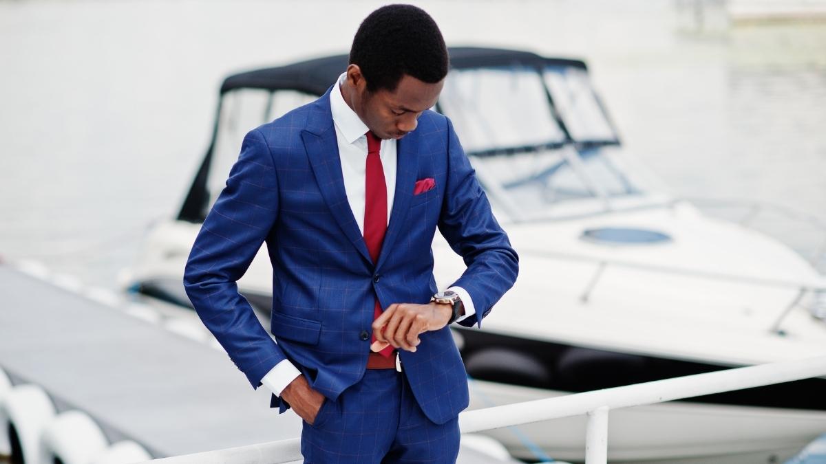 A well-dressed businessman in a blue suit checks his watch while standing in front of a yacht