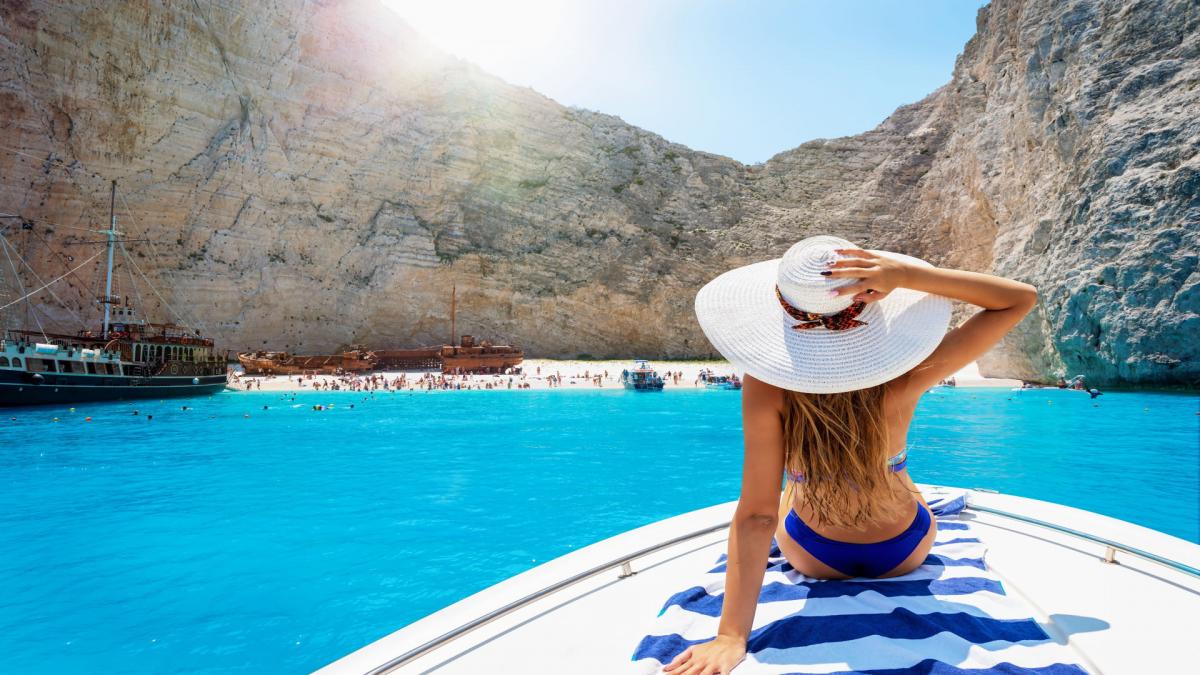 Rückansicht einer attraktiven Frau mit Sonnenhut im blauen Bikini die auf einer Yacht liegt und Bucht Anblick genießt.