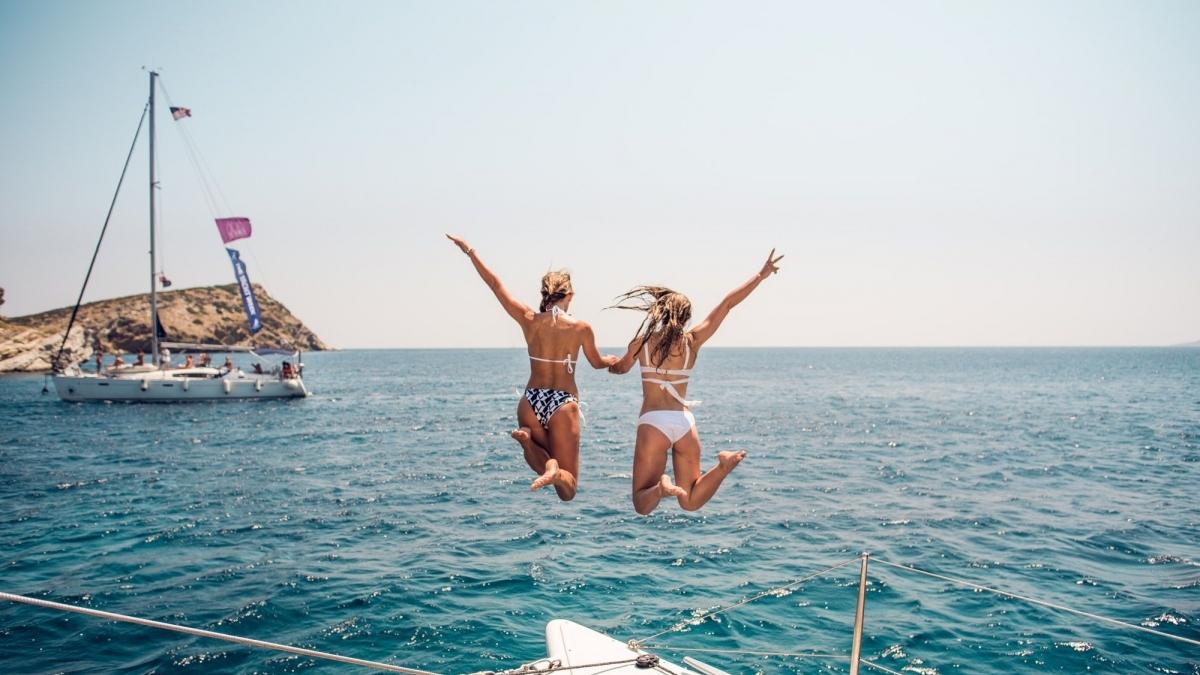 Two women joyfully jump from a yacht into the clear blue water, surrounded by sun and sea