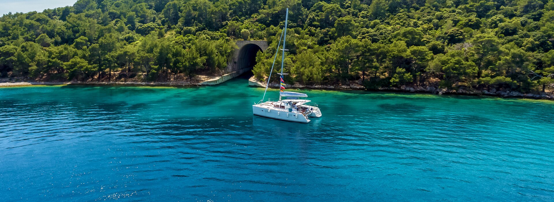 Белый катамаран в бирюзовой бухте у лесистого берега, аренда и чартер катамаранов в Салерно