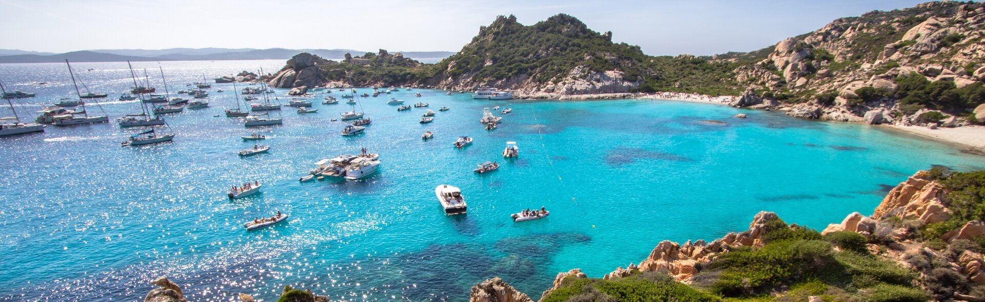 In einer türkisfarbenen Bucht auf Sardinien liegen zahlreiche Segelyachten und Boote vor Anker. Beliebtes Revier für Yac