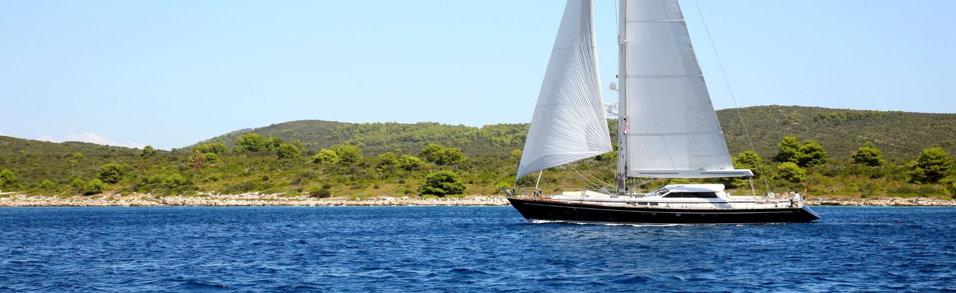 A stylish motorsailer yacht with full sails glides through the deep blue waters off the green coast of Lavrion