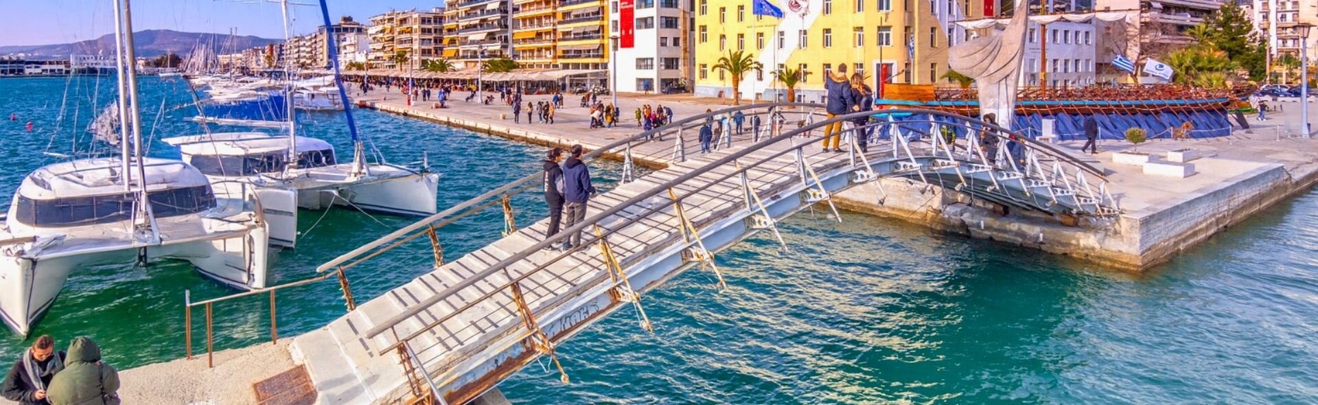 Blick auf die Hafenpromenade von Volos mit Katamaranen am Kai und der modernen Fußgängerbrücke unter blauem Himmel